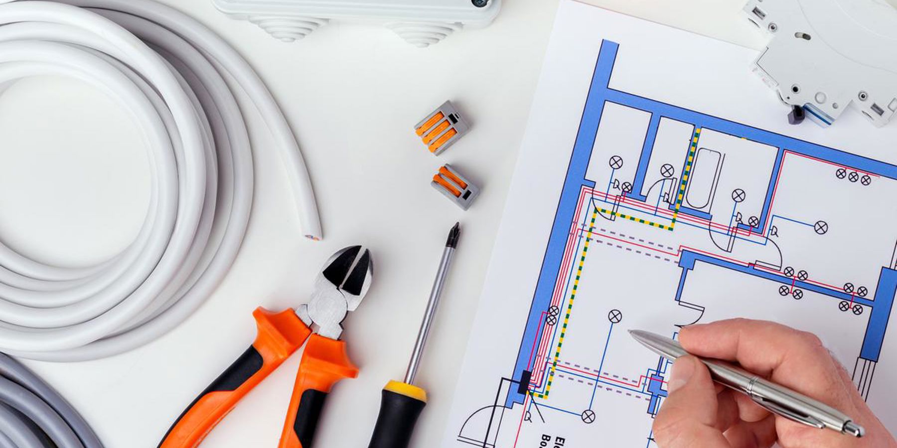 Hand holding pen over a building plan with electrical tools beside it.