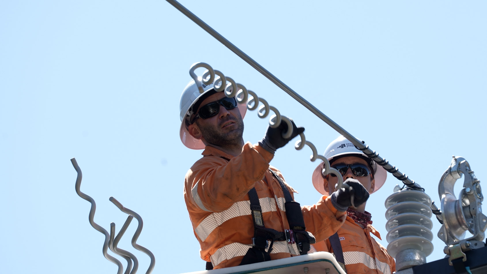 Two Western Power crew members installing a powerline covered with an insulating material an overhead power line from an elevated work platform.