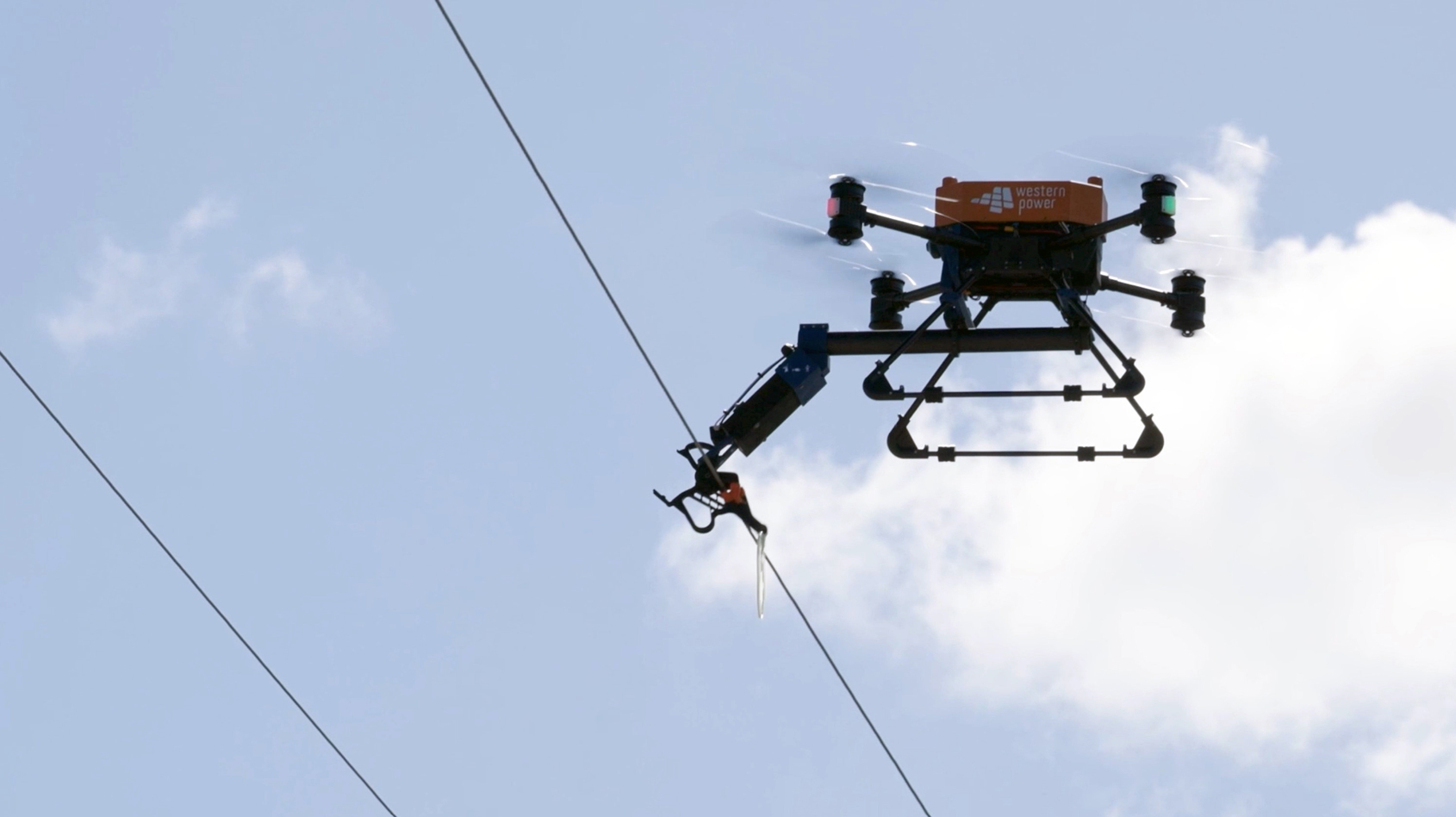 A Western Power drone flying near overhead power lines, using a robotic arm to work on the line.