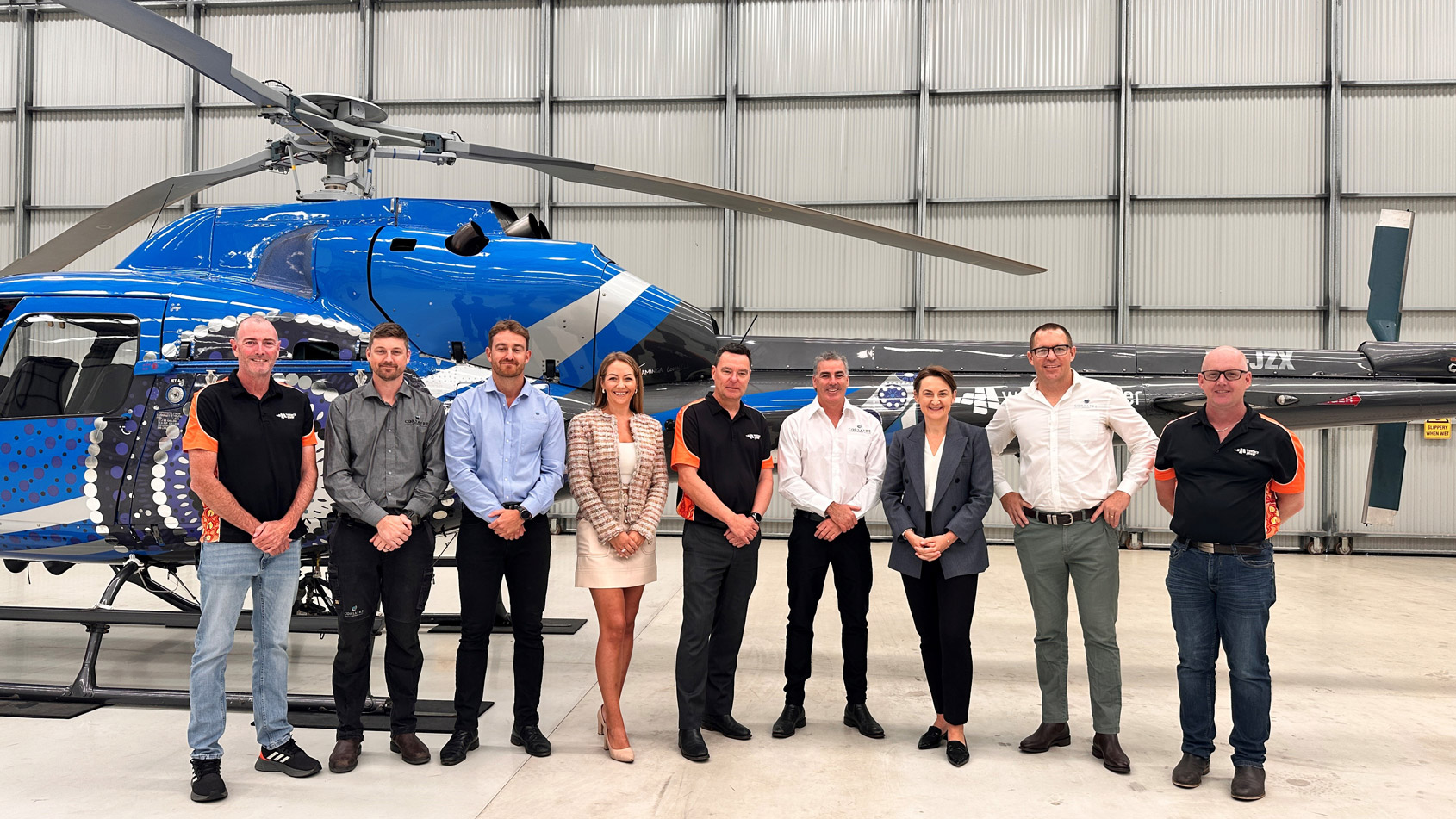 Nine adults, including Western Power employees and Hon. Amber-Jade Sanderson stand in a hangar in front of a blue helicopter, facing the camera and smiling.