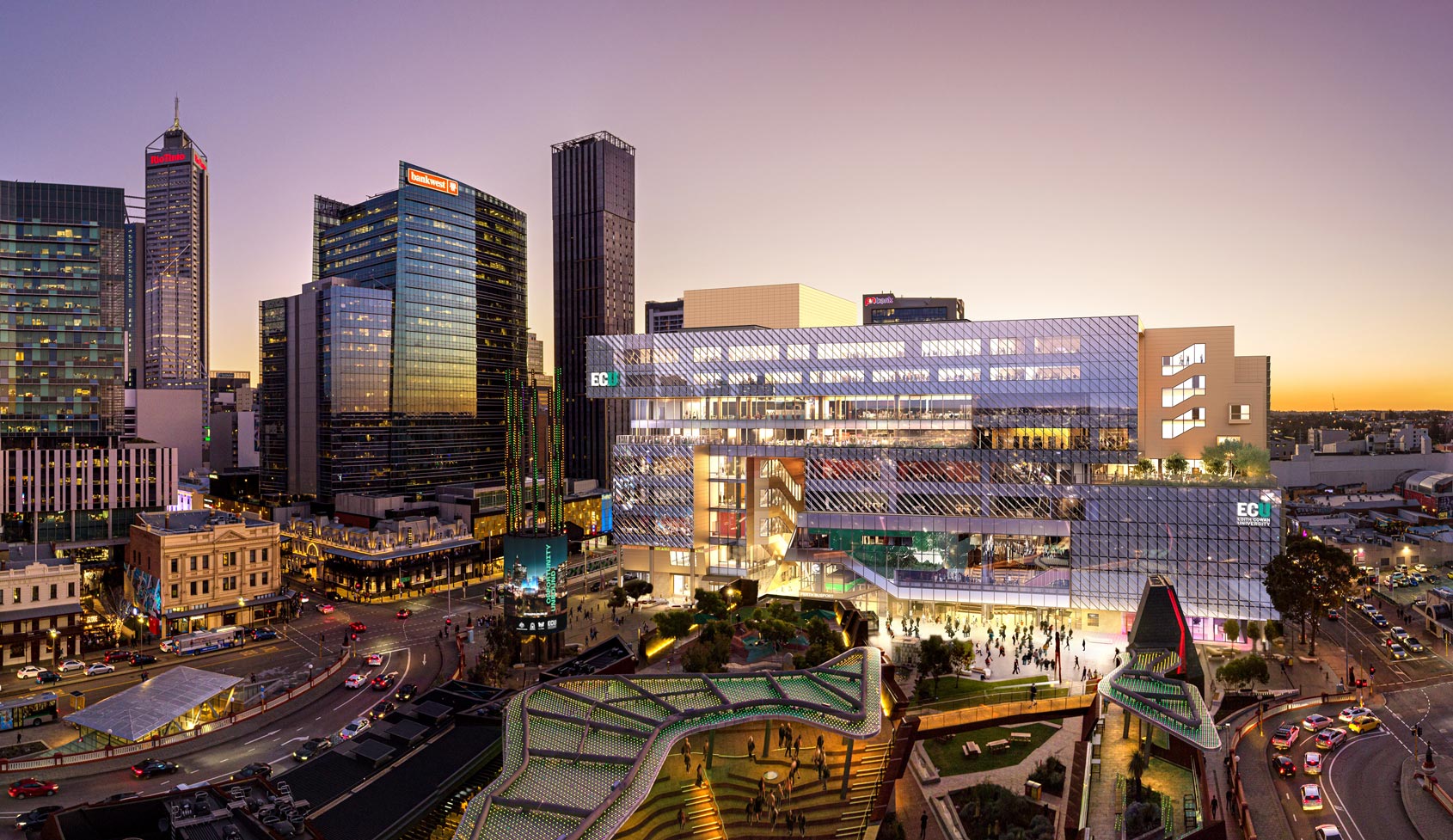Sunset view of the new ECU building with glass facades in a Perth city centre, surrounded by skyscrapers, streets with cars, and pedestrians in a lit plaza.