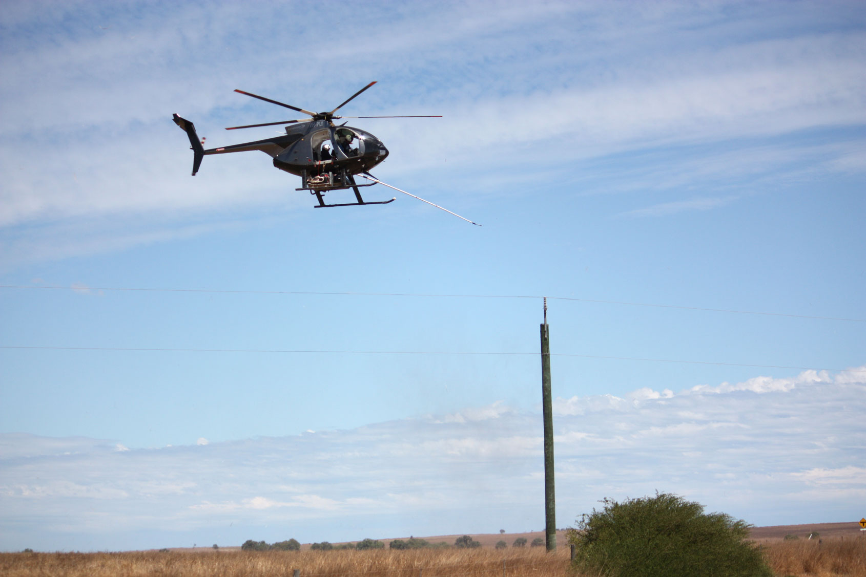 A helicopter hovers low over a rural field, carrying out line maintenance on overhead power lines.