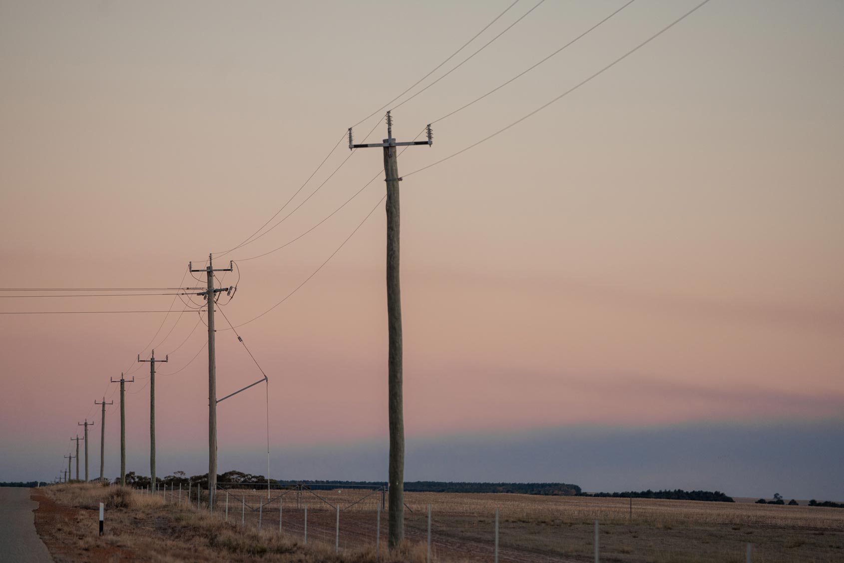 Power poles and overhead lines along a rural road at sunset, with open farmland beyond.