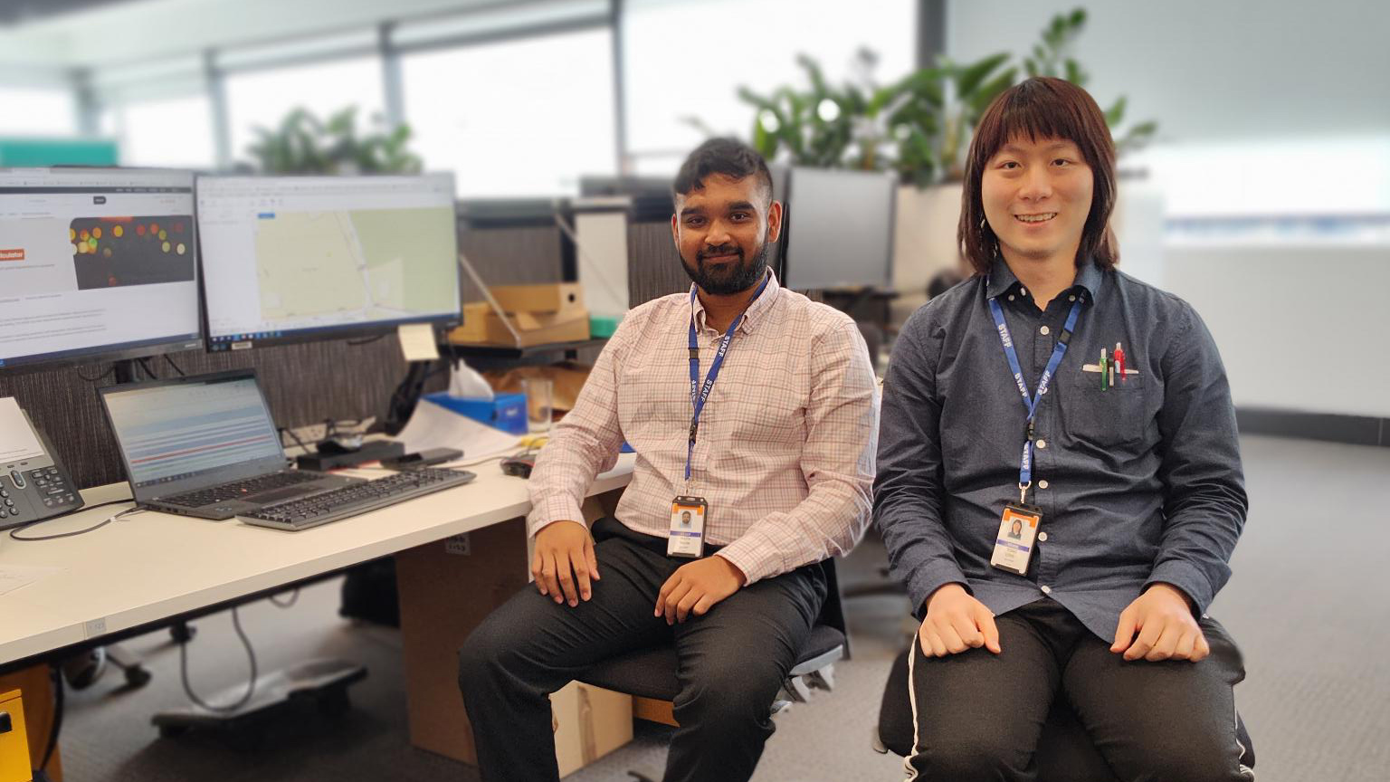 Two colleagues smiling at their desks in a modern office with computer screens behind them.
