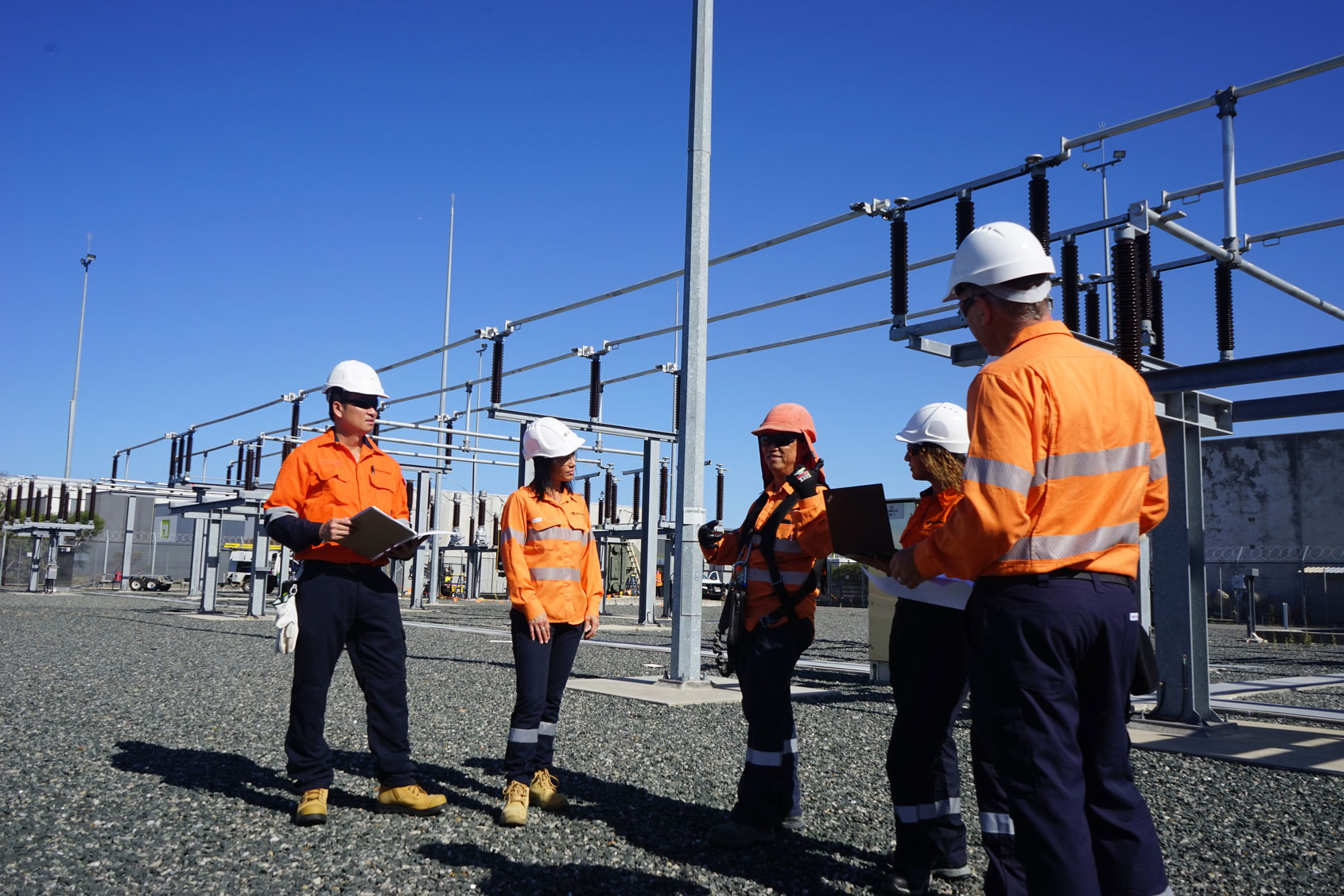 Workers in high-visibility orange safety clothing and hard hats conducting a site discussion at an electrical substation on a clear day.