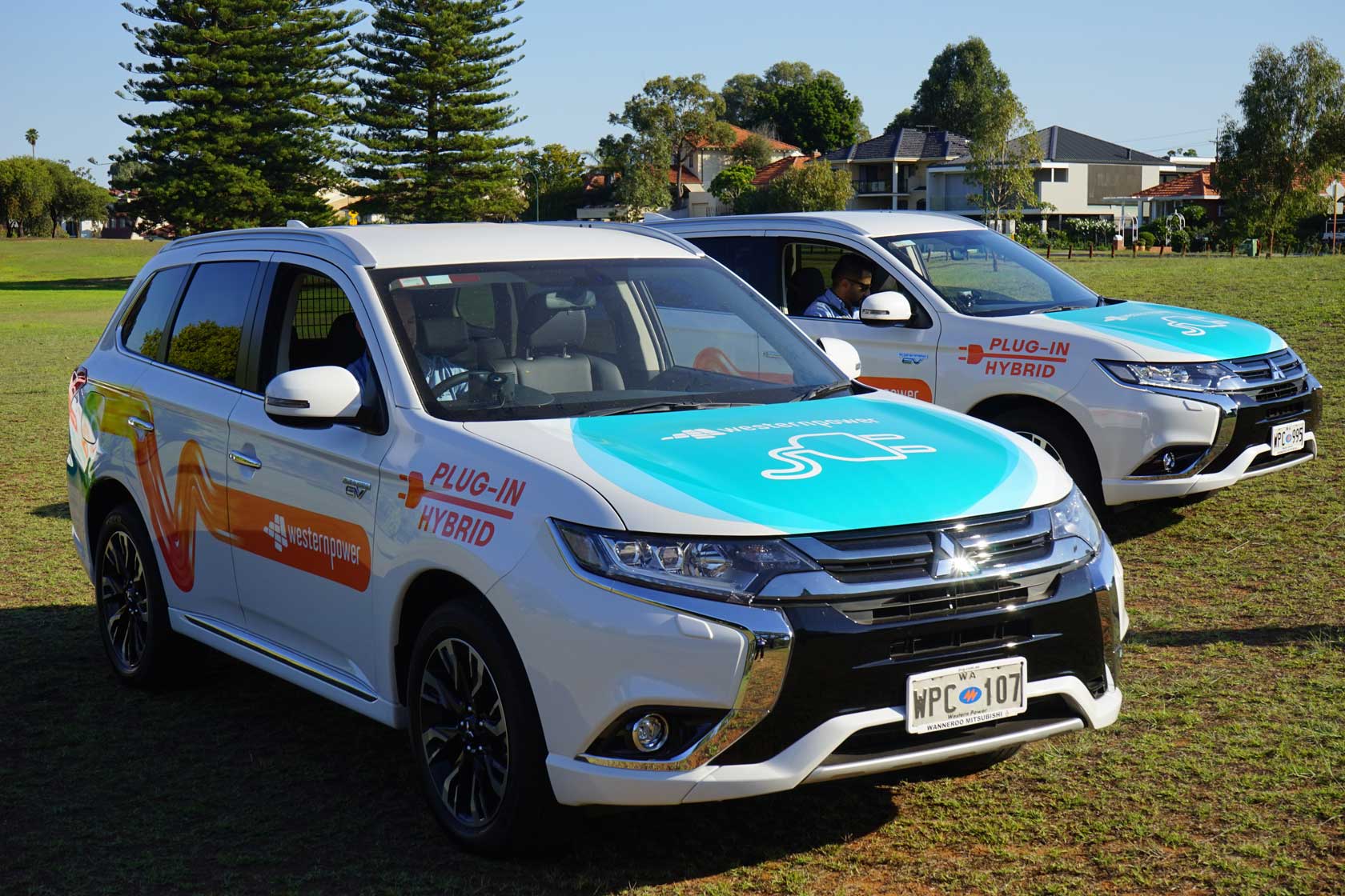 Two white Mitsubishi Outlander plug-in hybrid vehicles with Western Power branding parked on grass near trees and houses.