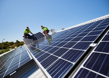 Two workers installing solar panels on a rooftop under a clear blue sky.