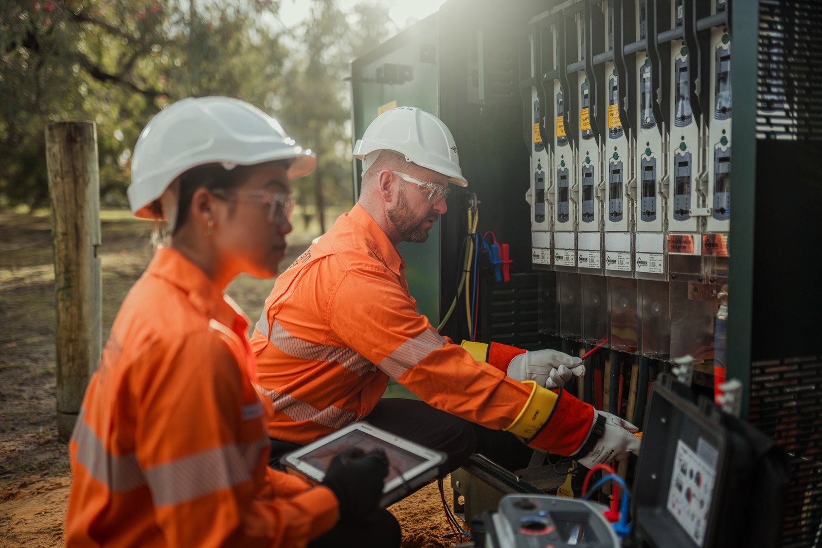 Two electrical workers wearing safety gear working on a distribution transformer. One worker is operating a testing device connected to the transformer, while the other appears to be observing or recording data on a tablet.