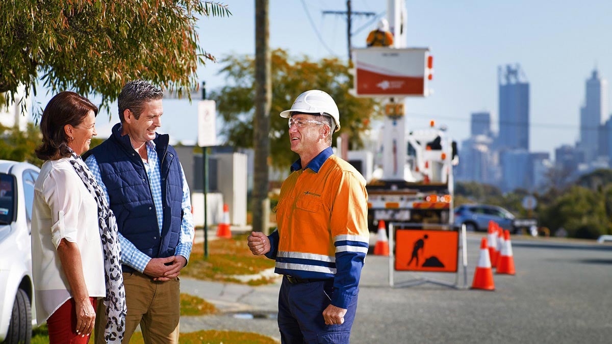 A Western Power crew member standing beside a road having a conversation with two members of the community. Traffic cones, construction signs, utility trucks, and equipment are visible in the background, with a city skyline in the distance.