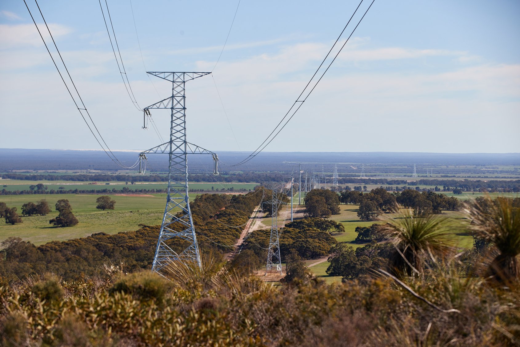 High-voltage power lines supported by transmission towers stretch across a green rural landscape under a clear blue sky.