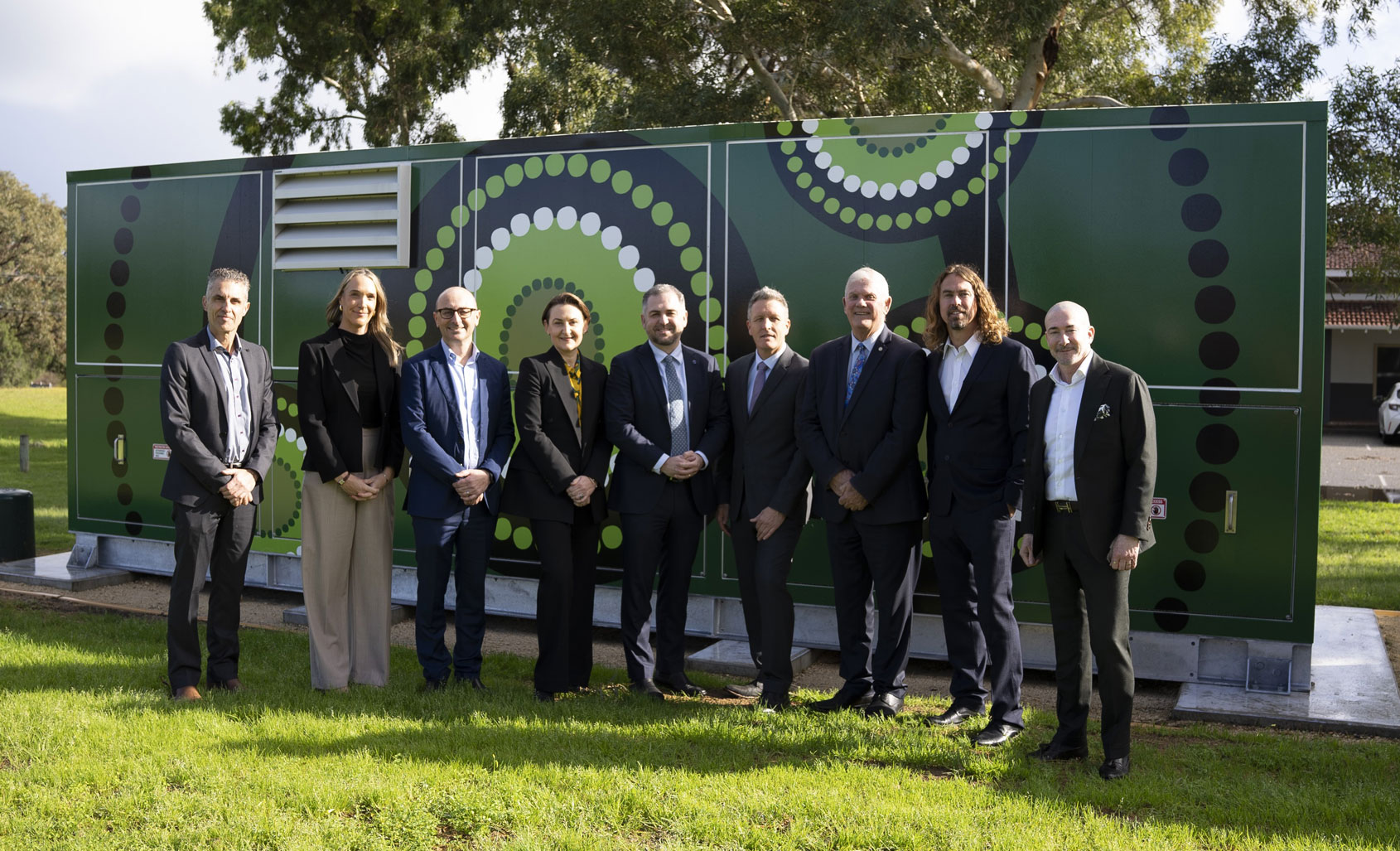A group of nine Western Power and Government representatives standing in front of a green community battery installation featuring Indigenous artwork, on a grassy area with trees and a building in the background.