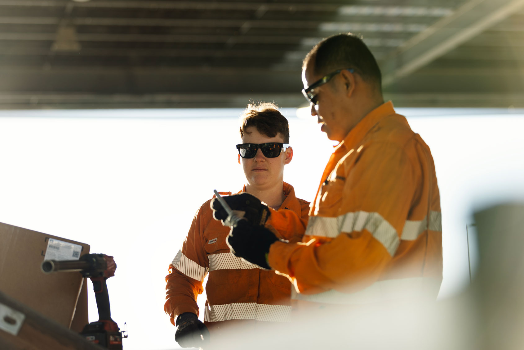 Two Western Power workers in orange safety gear examine a tool under bright daylight.