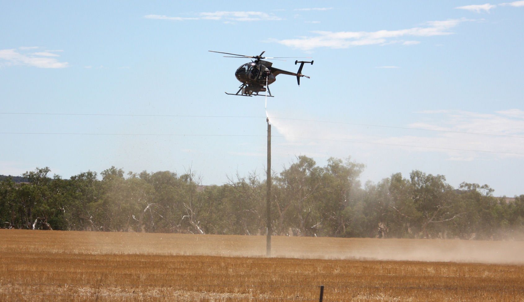 A helicopter spraying insulation or protective coating onto the top of a power pole in a rural field with trees in the background.