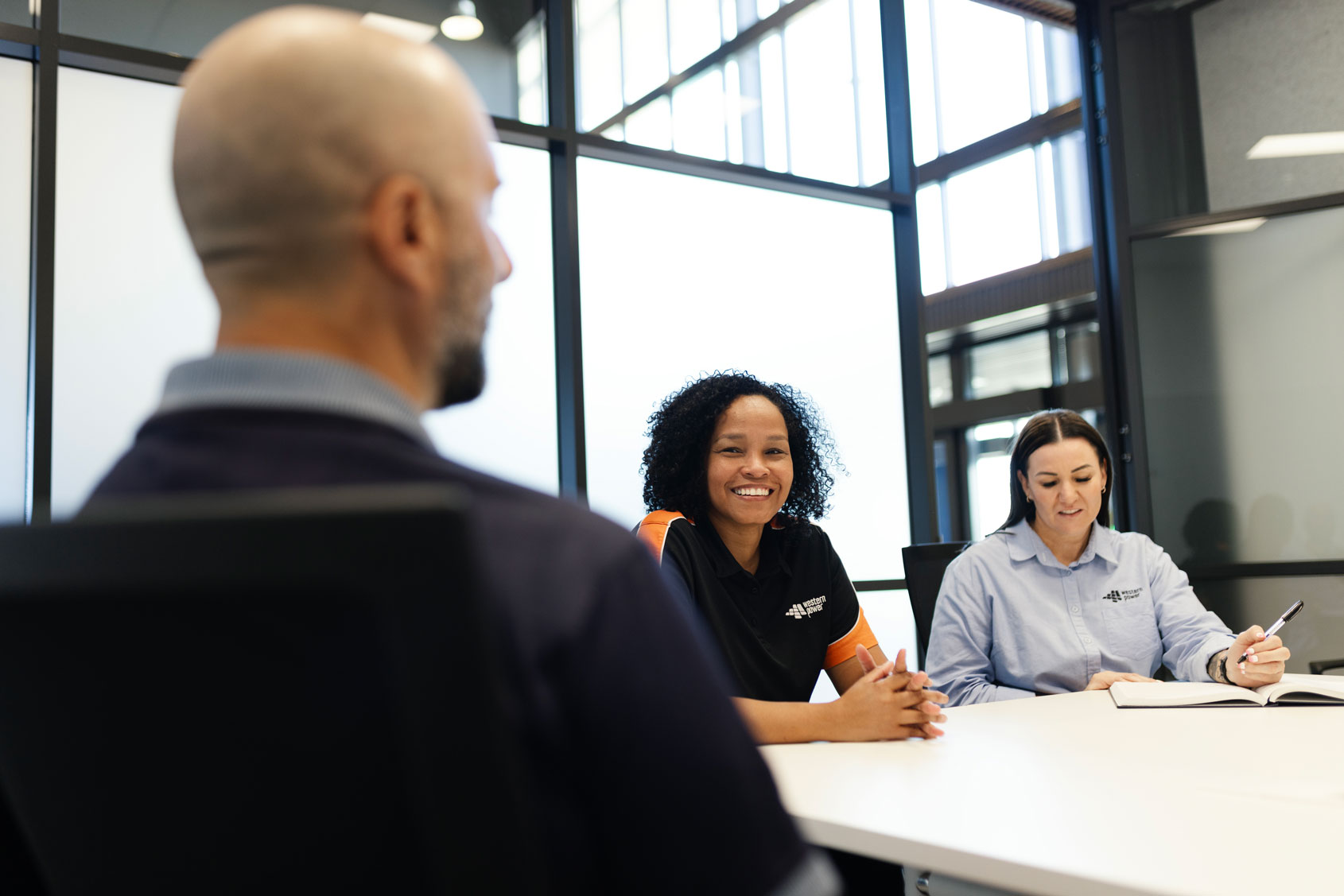 Three colleagues sit around a meeting table, engaged in discussion, with one woman smiling at the camera.