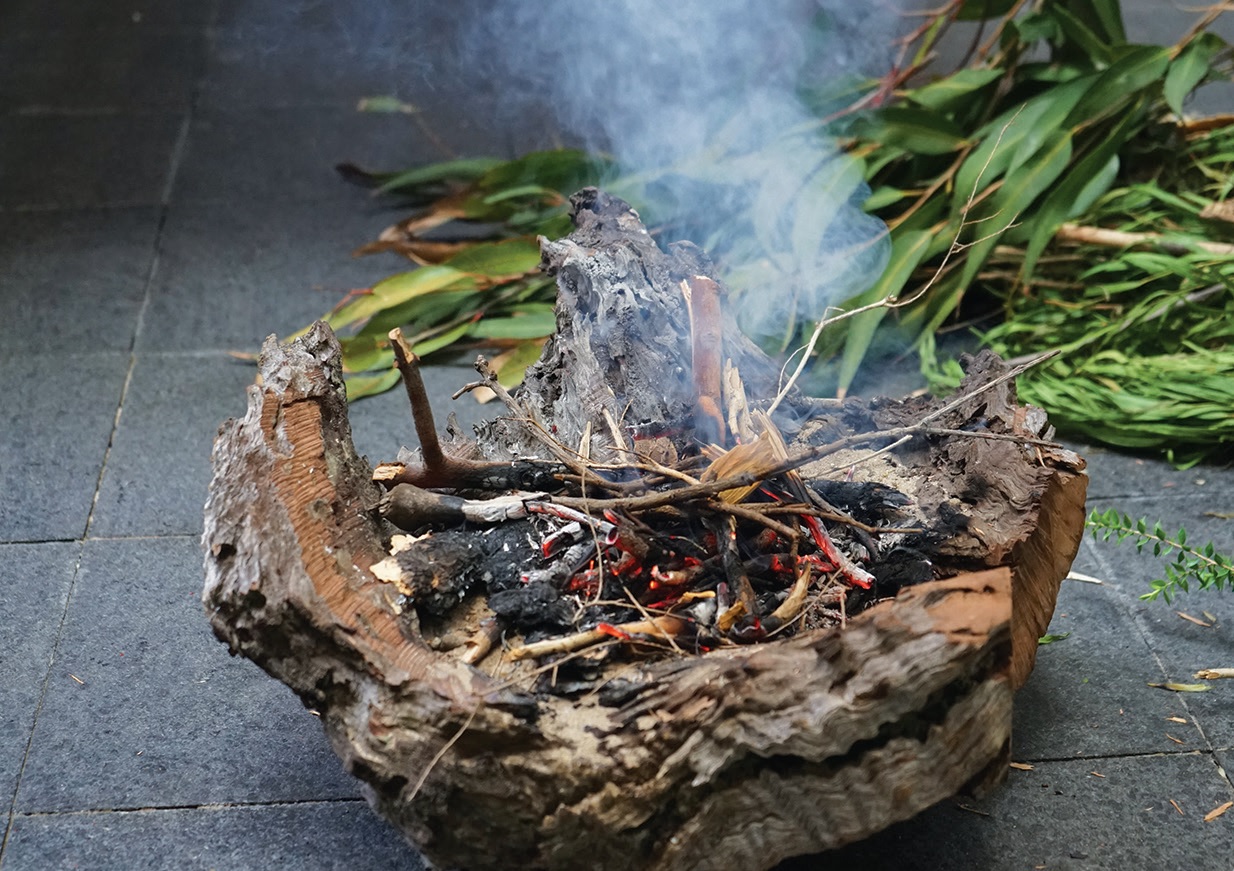 A smoking bundle of kindling and ashes sits in a bark container on a tiled floor, with green eucalyptus leaves nearby, creating a natural, earthy scene.