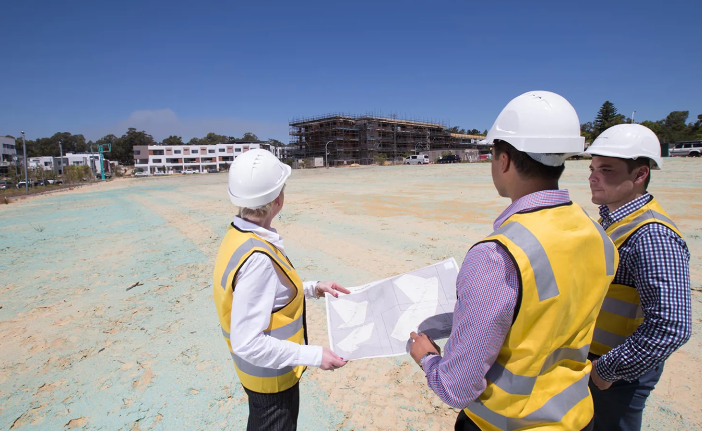 Three construction workers wearing safety vests and helmets examine blueprints on a large, empty construction site, with a partially built structure in the background.