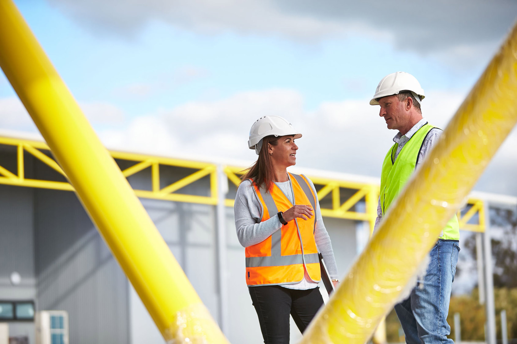 Two construction workers wearing hard hats and bright vests converse outdoors near a building with yellow beams, under a partly cloudy sky.