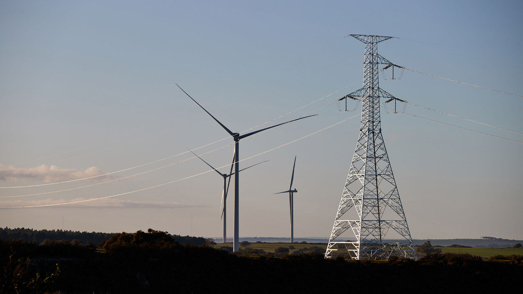 ind turbines and a transmission tower with power lines in a rural landscape under a clear sky.