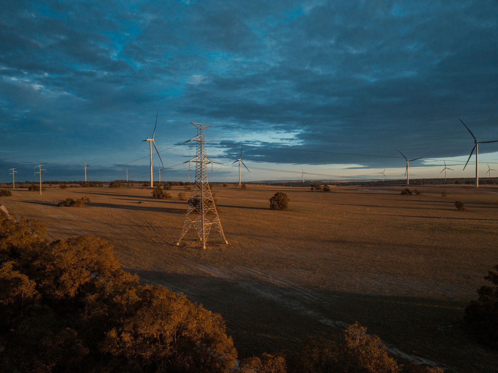 Transmission towers and wind turbines in a rural landscape at sunset, with power lines stretching across open fields.