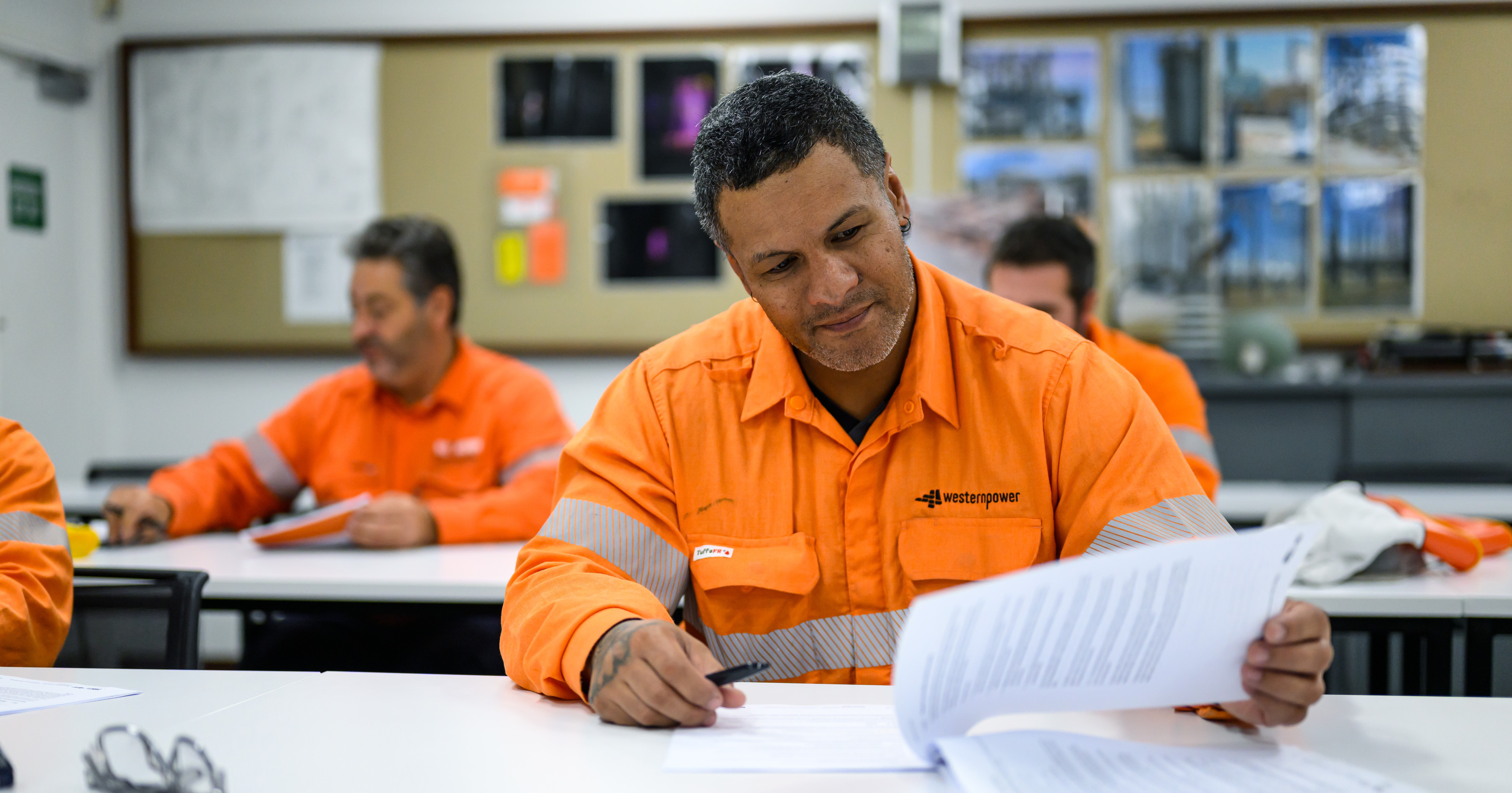 A group of men in orange PPE seated at tables in a classroom-styled environment, looking through documents.