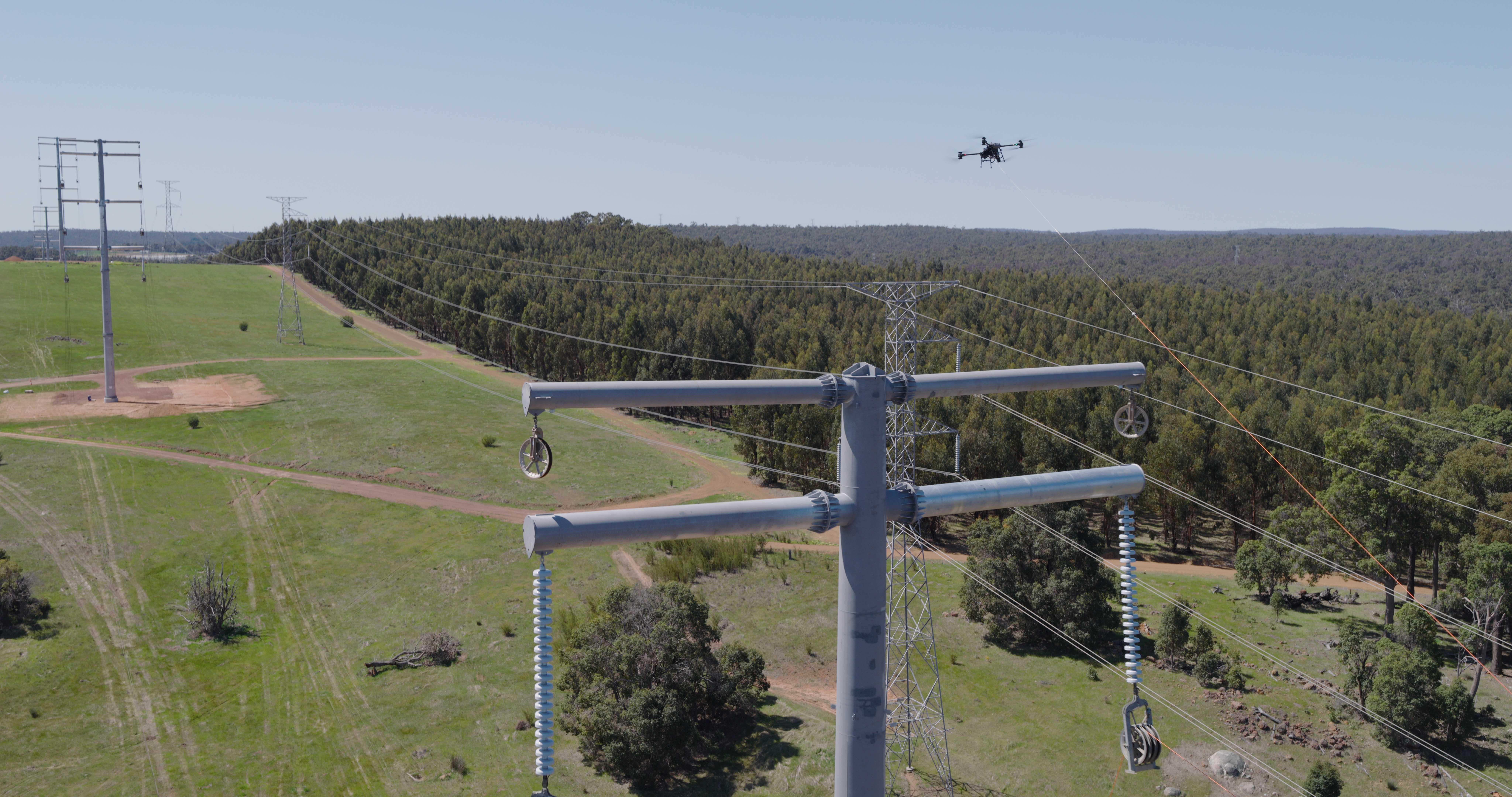 Transmission towers with forest in the background and a drone flying overhead with stringing a powerline.