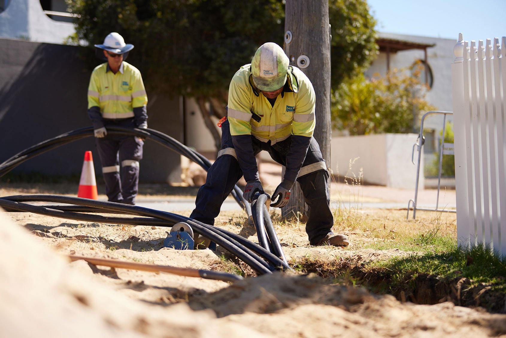 Two utility workers in high-visibility clothing installing underground cables on a suburban street.
