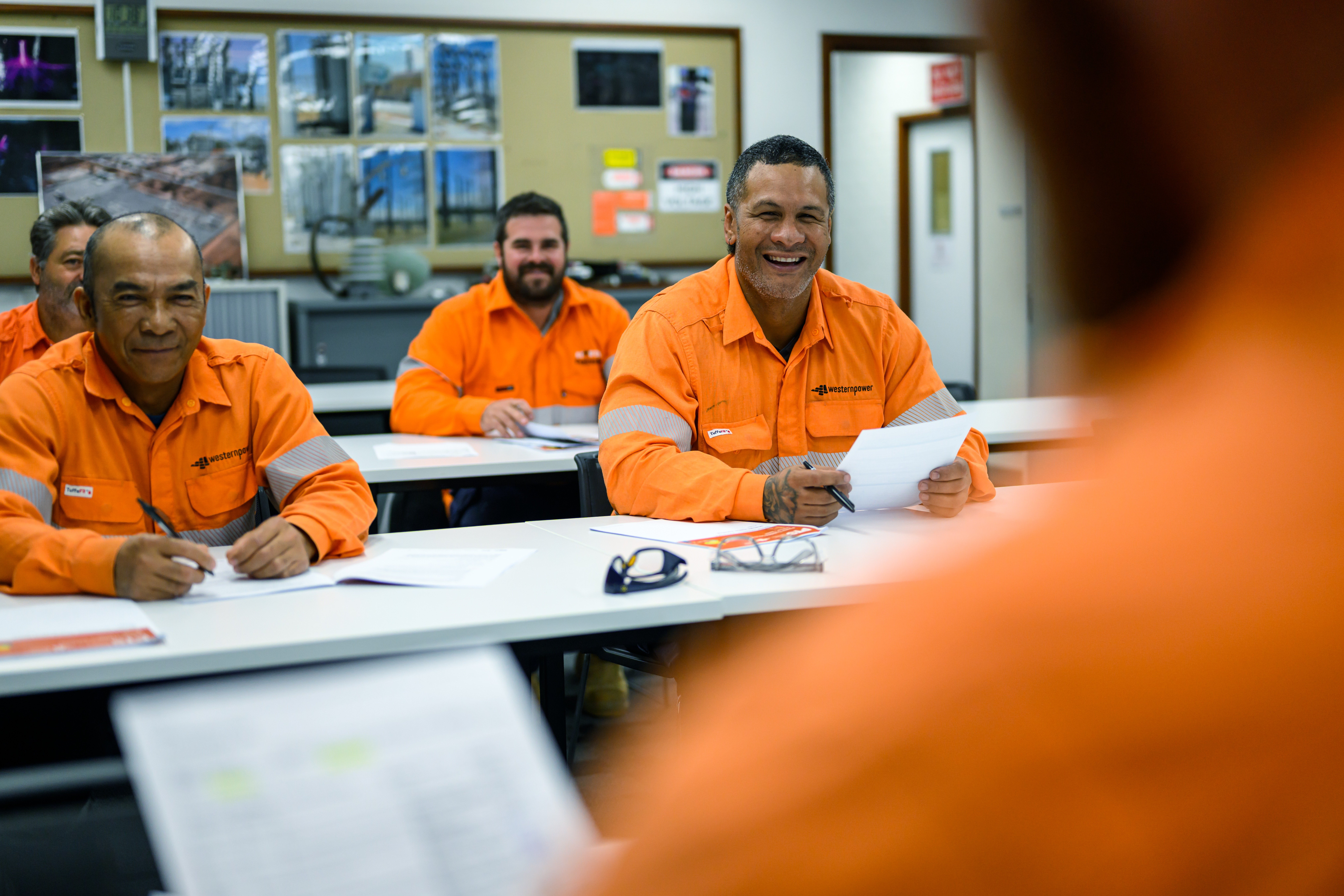 Electrical safety training session with workers in orange PPE seated at desks, reviewing documents and notes in a classroom setting.