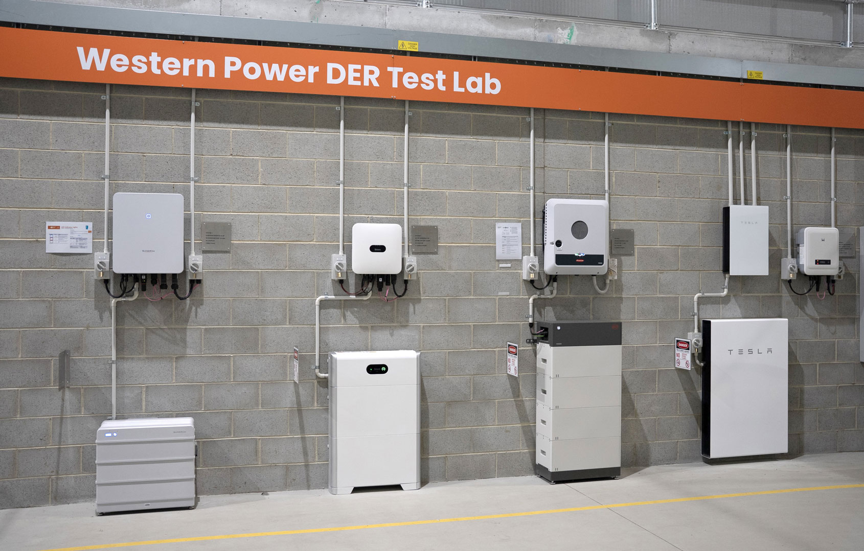 Wall in Western Power DER Test Lab with multiple inverters and battery systems mounted on a grey brick wall under an orange sign.