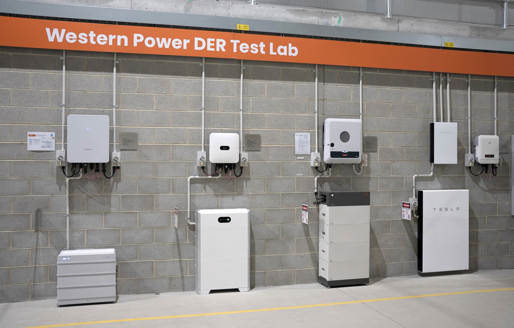 Wall in Western Power DER Test Lab with multiple inverters and battery systems mounted on a grey brick wall under an orange sign.