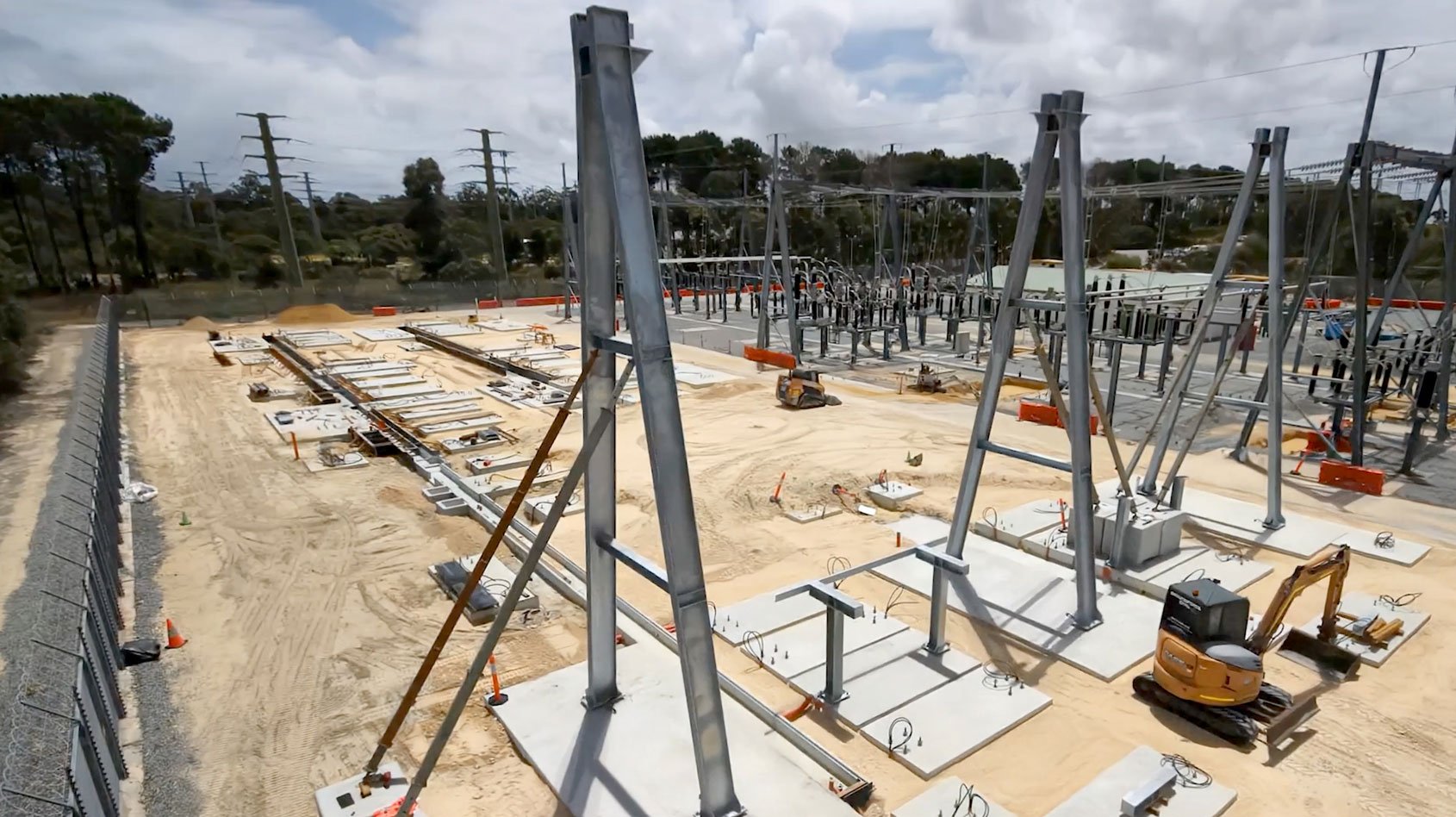 A construction site with large steel framework structures being installed on concrete foundations, surrounded by sand, machinery, and existing electrical substation equipment in the background.